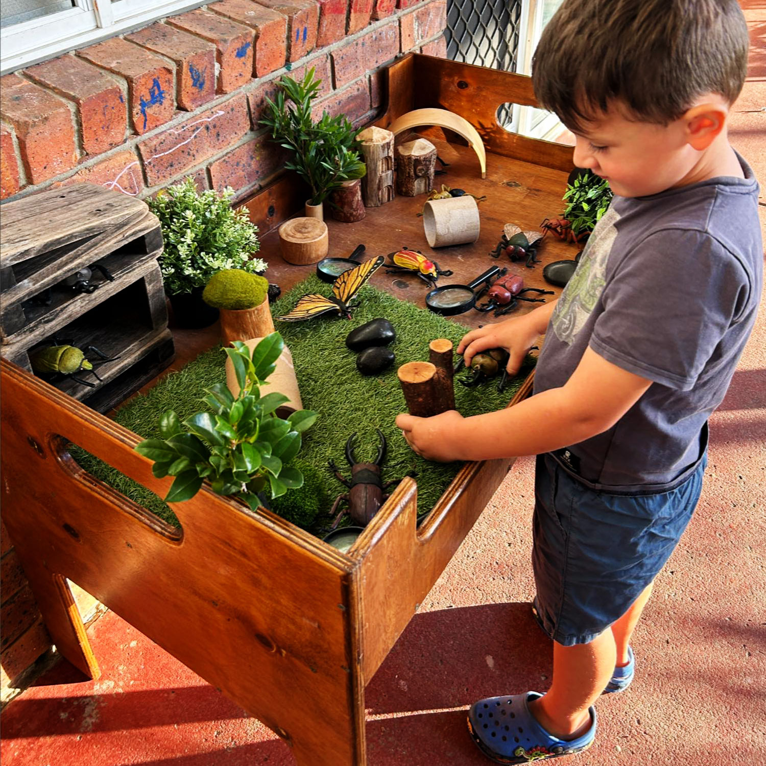 A photograph of a young child playing at a wooden table that is partly covered with fake grass. On the table there are plants and wooden pieces along with a range of insect toys.