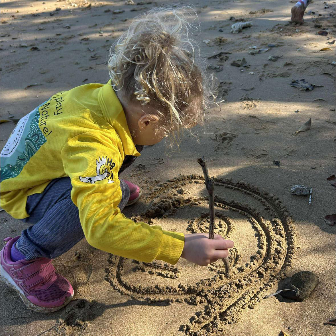 A photograph of a child drawing a picture of a face in the sand with a stick.